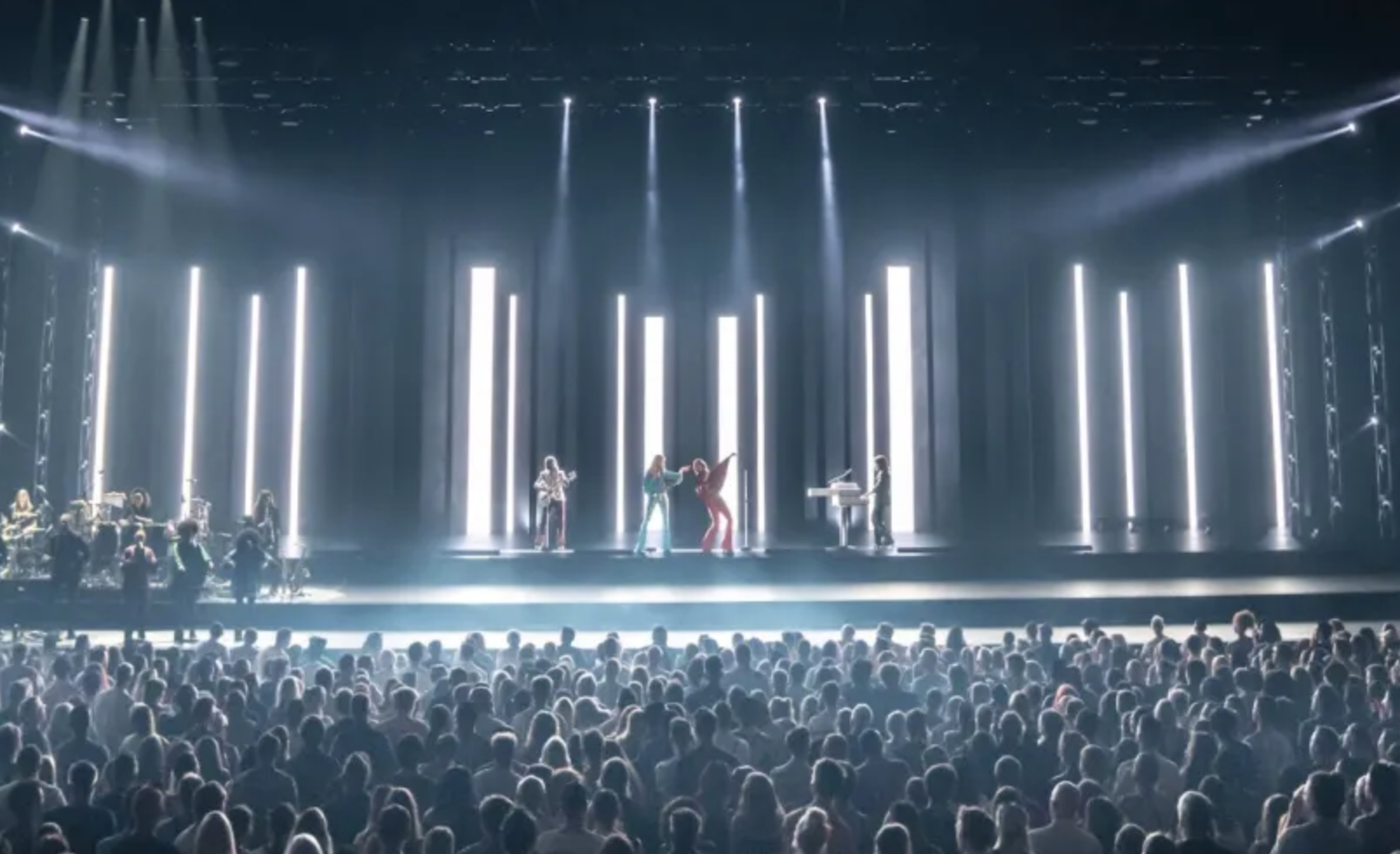 Band performing on a stage with vertical light panels; large audience watching the illuminated show in a dark venue.