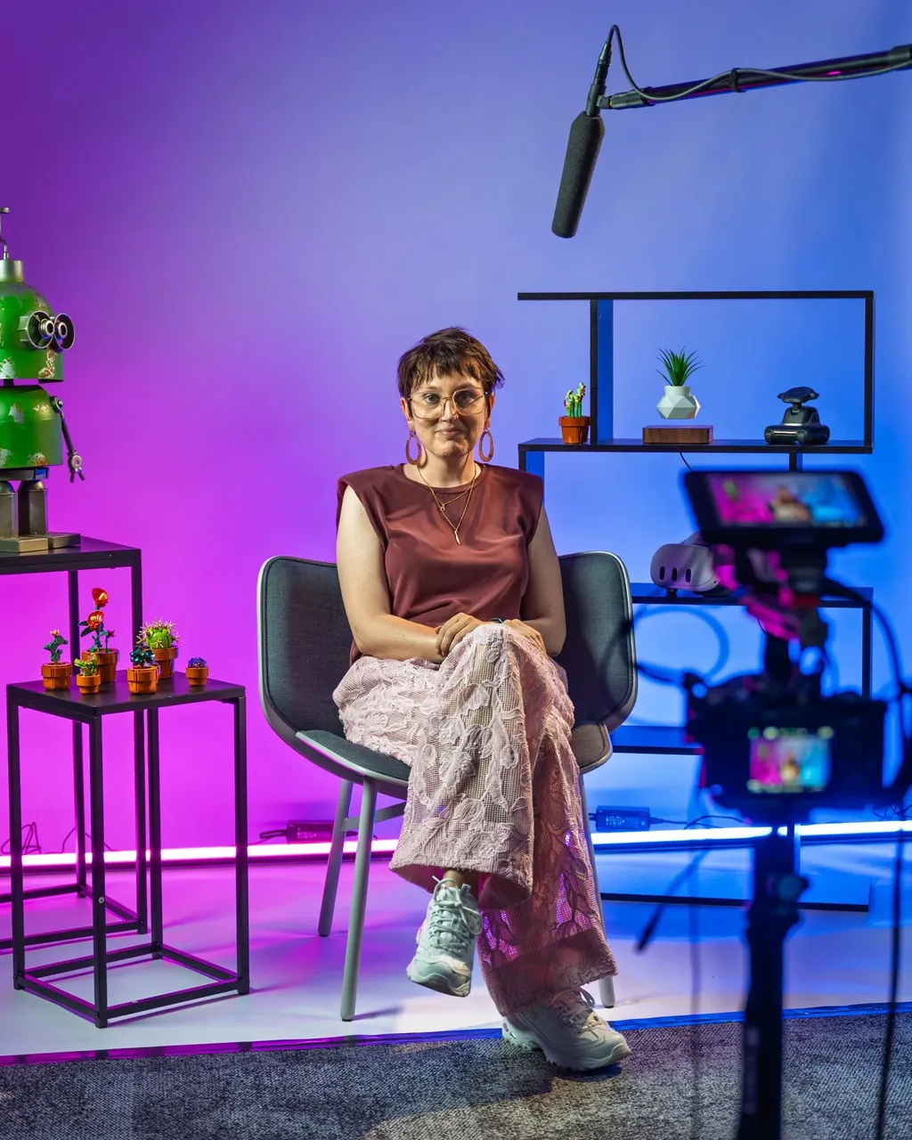 Ine Vanoeveren, woman,  sits on a chair in a colorful studio with plants and a camera setup, wearing glasses and stylish attire.