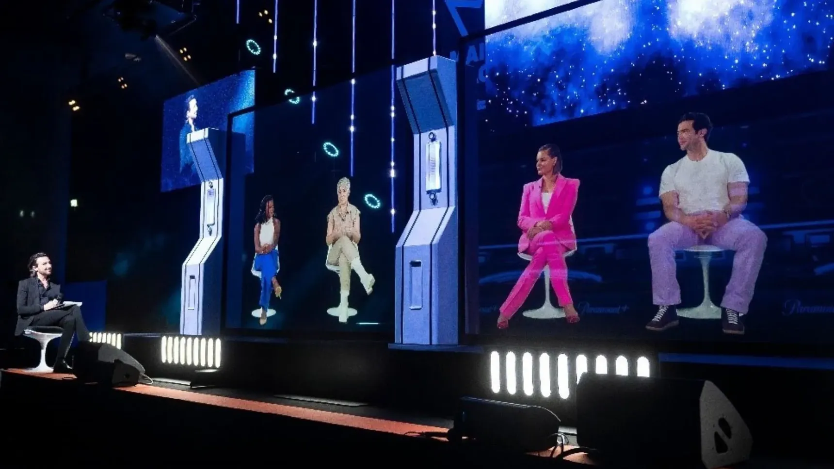 A person moderates a panel with virtual images of four individuals on a stage with digital screens and vibrant lighting effects.