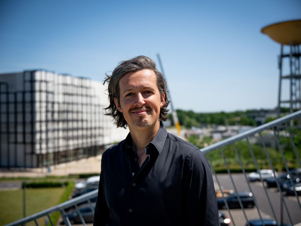 Man in a black shirt stands outdoors on a sunny day, with a modern building and a construction site in the background.