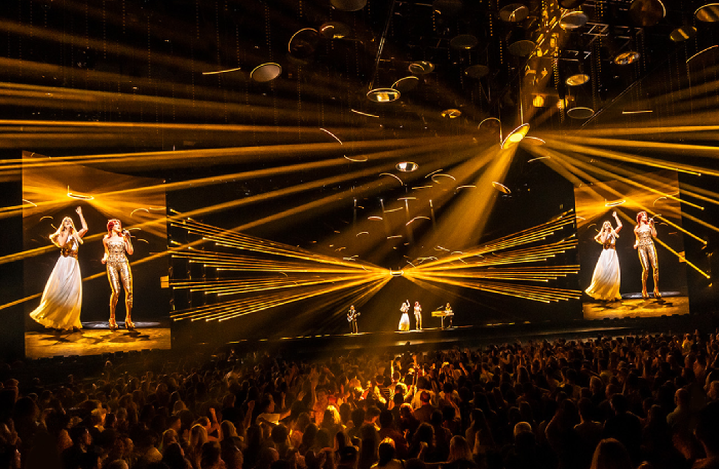 A lively concert scene with a band performing under vibrant golden lights, flanked by large screens, in front of a cheering crowd.