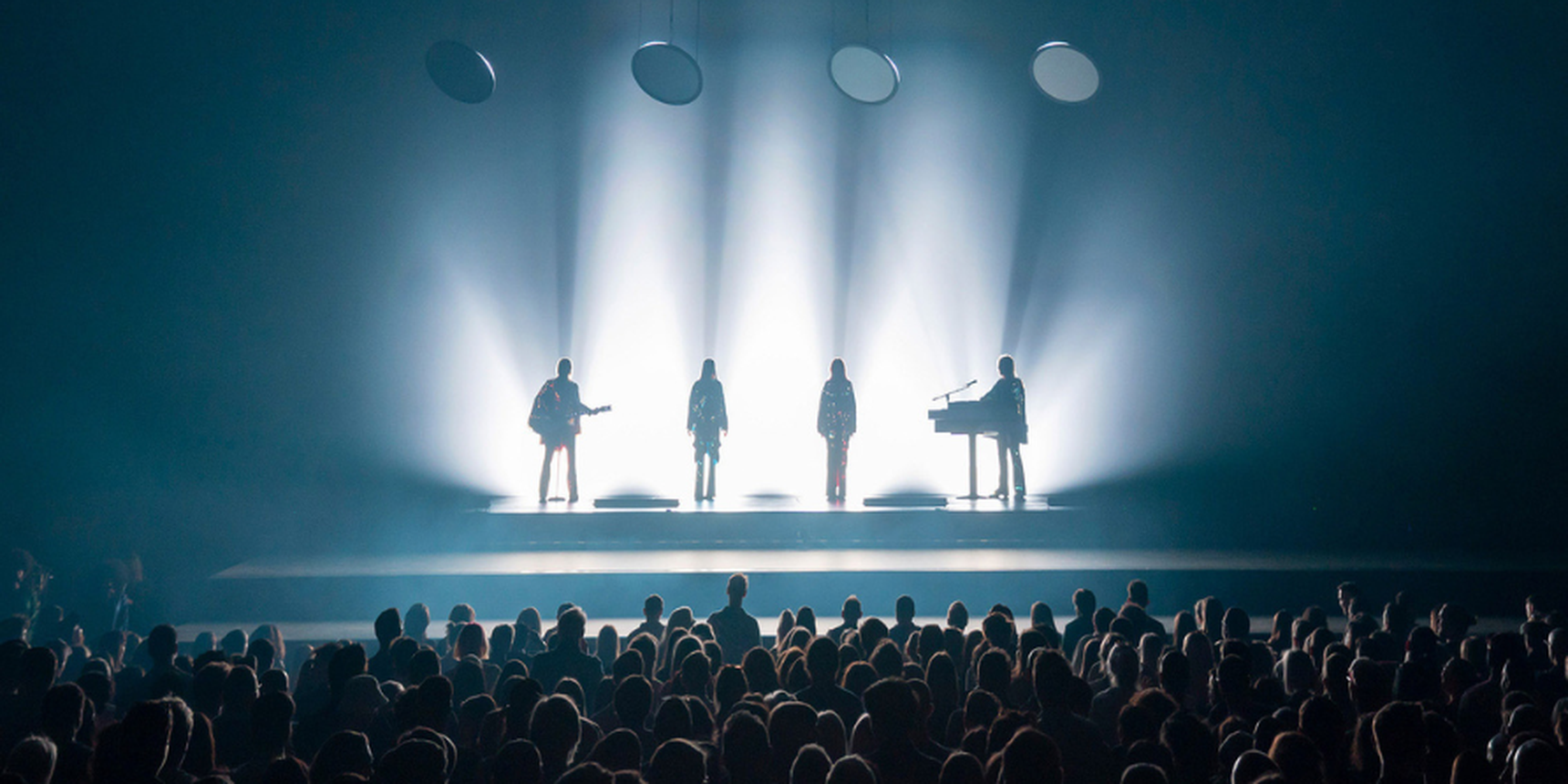 Silhouetted band performs on stage with bright backlighting, facing a captivated audience.