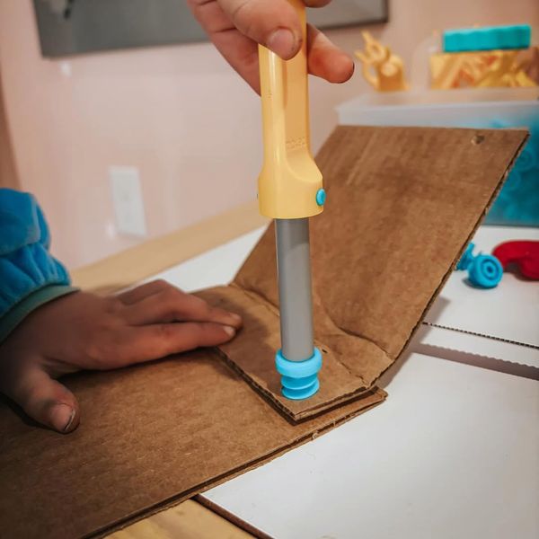 A child uses a Makedo Scru-Driver to connect two pieces of cardboard with a bright blue Makedo Scru connector.