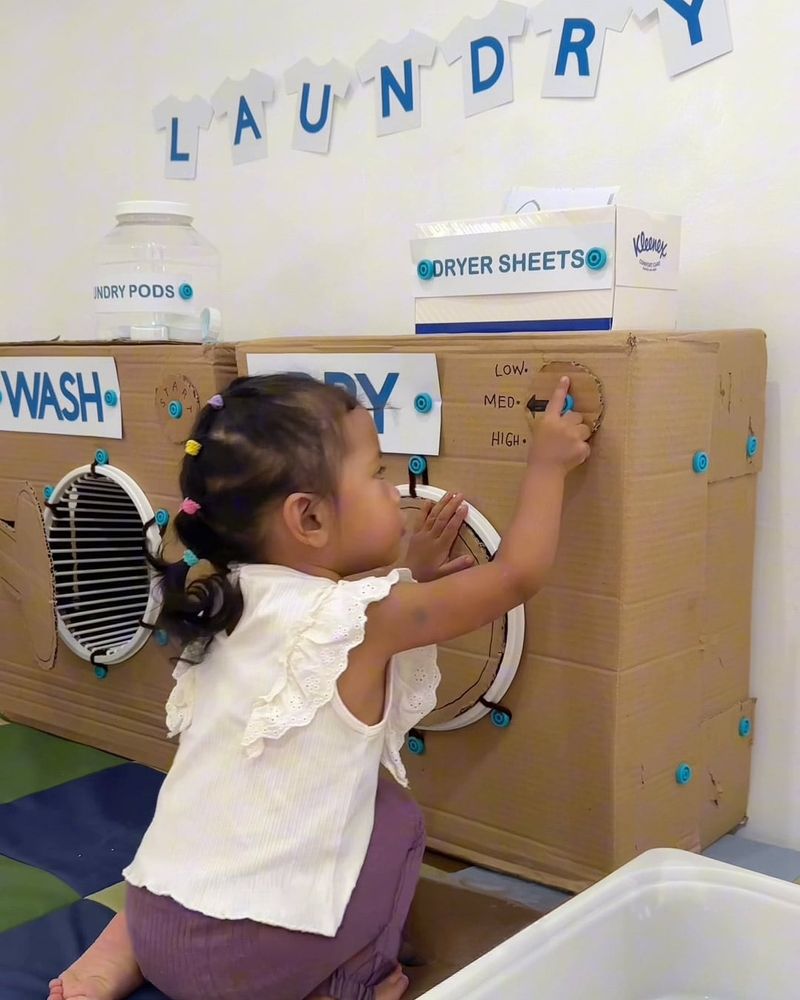 Child playing with a cardboard Makedo laundry, adjusting dials. Wall letters spell "LAUNDRY."