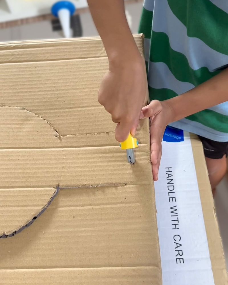 Child marking a cutting line in cardboard with a yellow handled Makedo Fold-Roller cardboard construction tool.