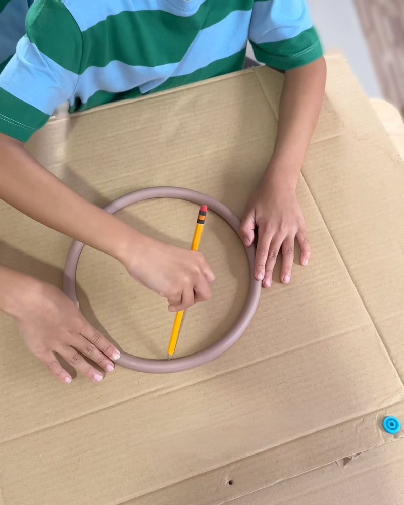 Child tracing a circle on cardboard using a plastic ring and pencil, wearing a green striped shirt.