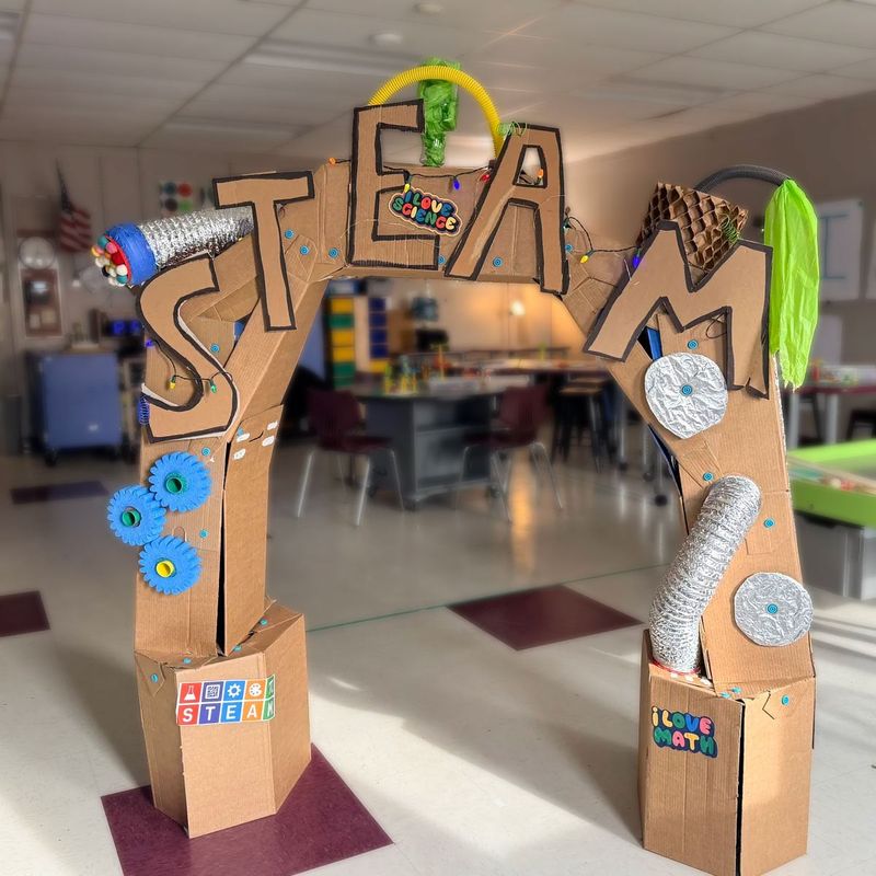 Cardboard archway constructed using Makedo and labeled "STEAM," decorated with gears and metal tubes, set in a classroom with tables and chairs in the background.