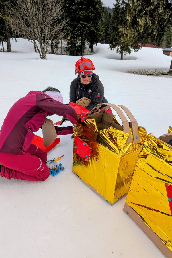 Two adults working on a cardboard Makedo dragon costume in the snow. They have Makedo cardboard construction tools in a ziplock bag.