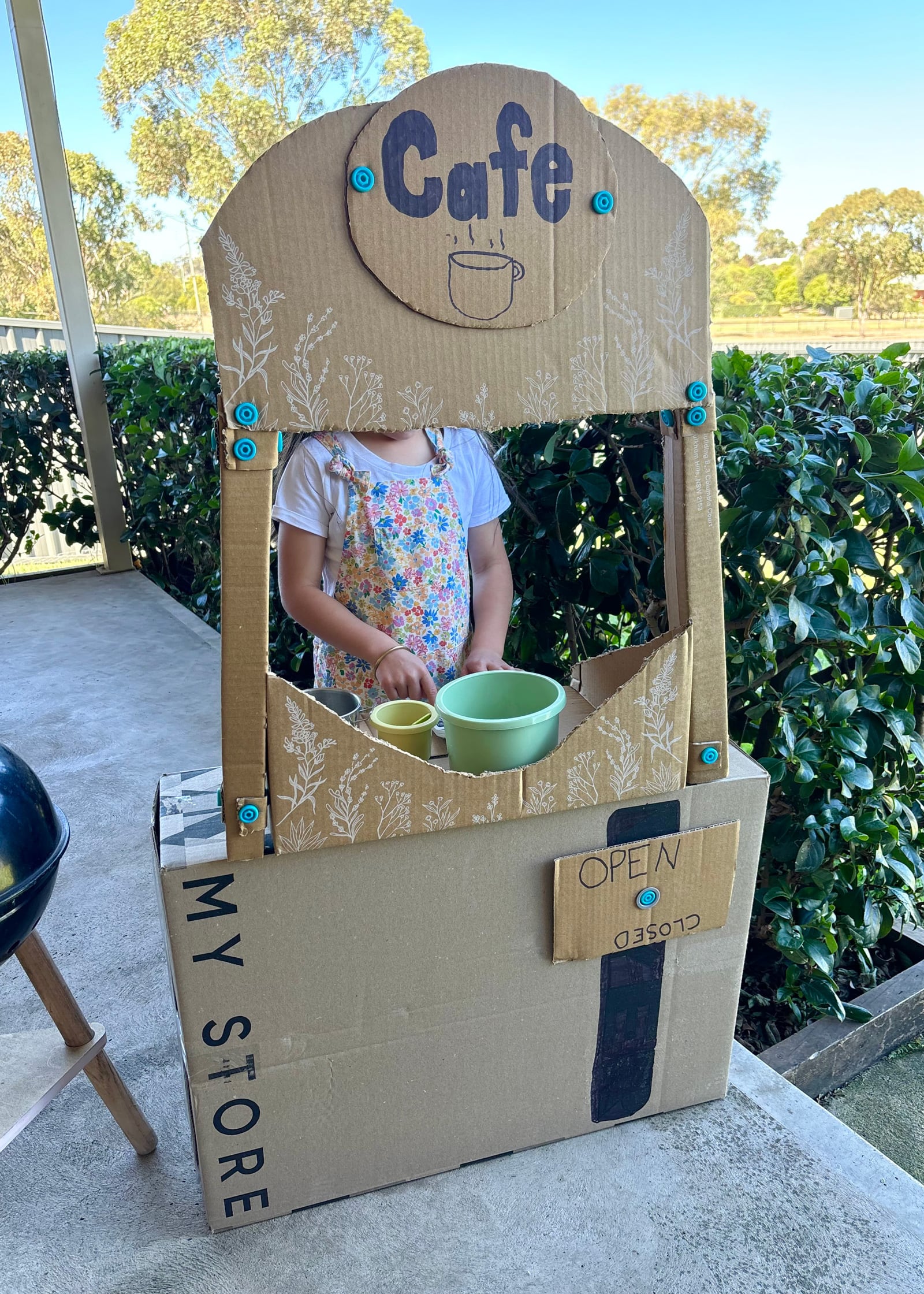 A child stands at a cardboard Makedo cafe stall, there is a rotating Open/Closed sign on the front.