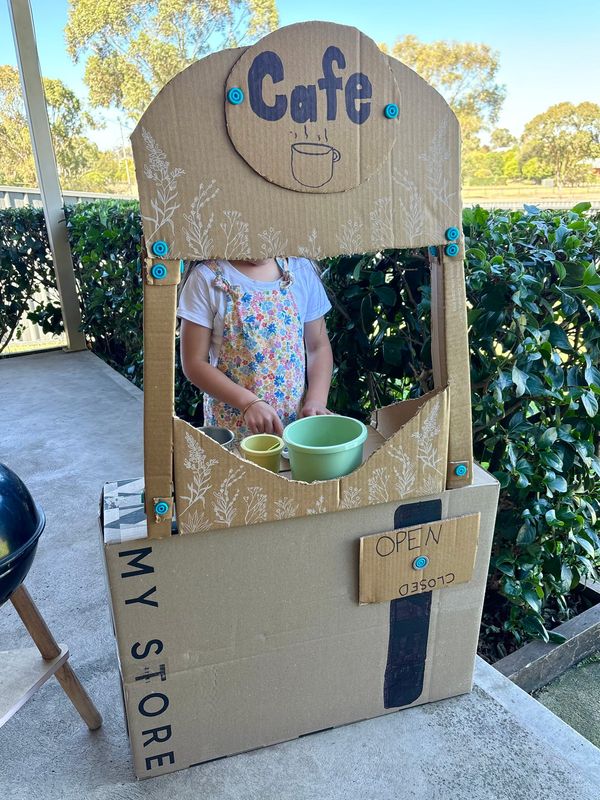 Child standing behind a play cafe that they have made using cardboard and Makedo. The cafe is marked "Open" on a rotating sign.