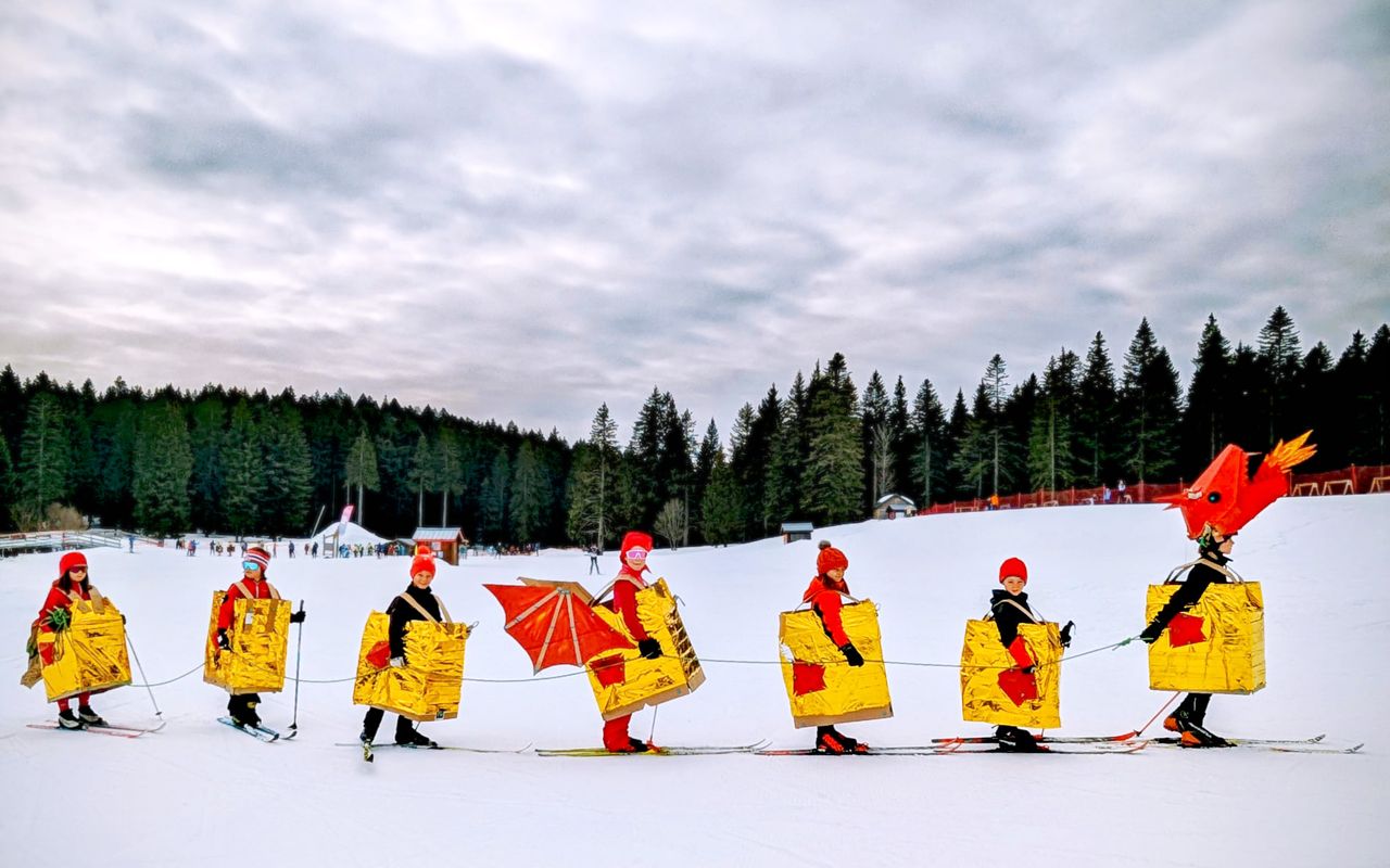 Group of skiers wearing a group dragon costume made from cardboard and Makedo, glide across a snowy landscape with evergreen trees in the background.
