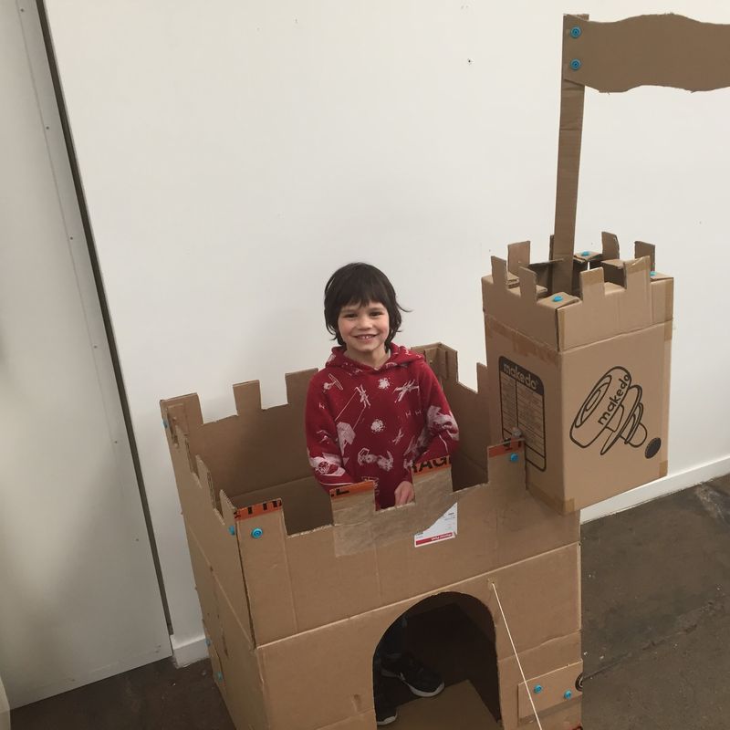 Young boy wearing a red top proudly standing inside a cardboard castle built with Makedo