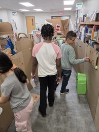 Children walking through a maze made of cardboard boxes in a classroom, with bookshelves and light gray flooring.