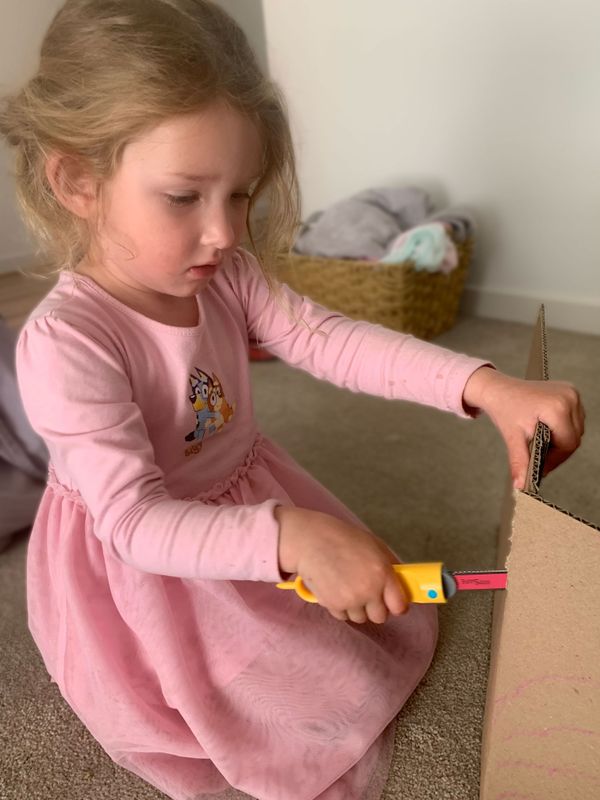 Young girl in a pink dress sitting on the floor using a Makedo Safe-Saw to cut corrugated cardboard.
