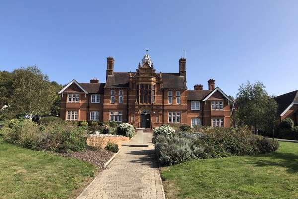 Ornate red brick building with a central bay, large windows, and a cupola, surrounded by green gardens and a paved path under a clear blue sky.