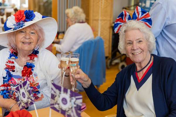 Two smiling elderly women celebrate. One wears a white hat with red & blue flowers and a lei, the other a Union Jack headband