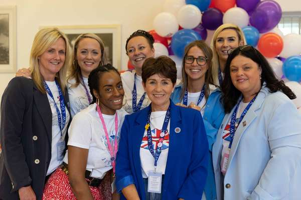 Eight smiling women wearing New Look t-shirts and blazers pose in front of red, white, blue, and purple balloons.