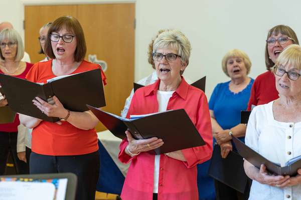 A choir of women, mostly older, sing with open mouths, holding music folders. They wear casual clothing, some with glasses.