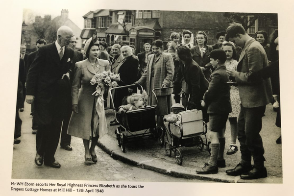 Queen Elizabeth, in a coat and hat, holds flowers while escorted by a man past a crowd, prams, and a red phone booth in 1948.