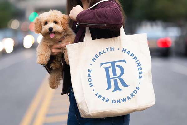 A person holds a fluffy tan puppy and an off-white tote bag with a blue "TR" logo and text "HOPE HEALTH HAPPINESS 1832 ONWARDS" on a street.