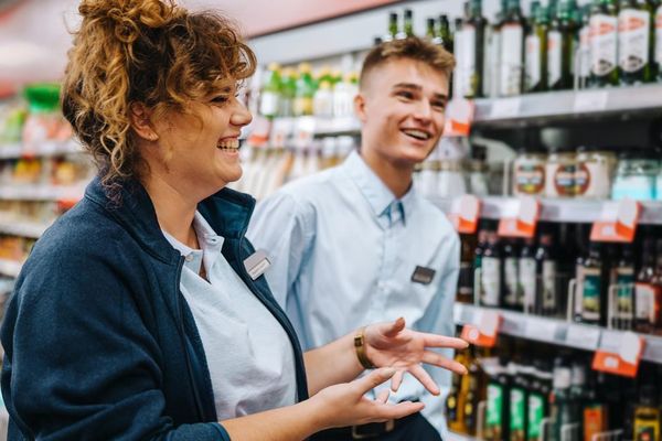 Two smiling supermarket employees, a woman and a young man, stand in an aisle with shelves of products. The woman is gesturin