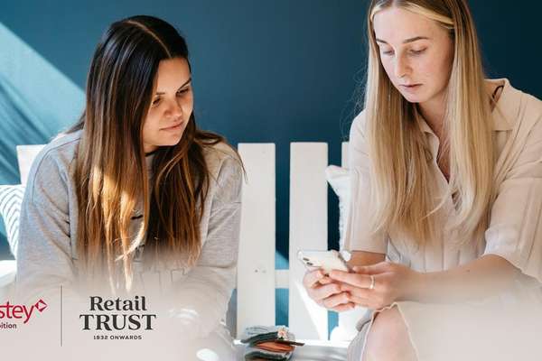 Two young women relax indoors. One uses a phone, the other observes. Blue wall with sunlight shadows, white furniture. FootAnstey and Retail Trust logos.