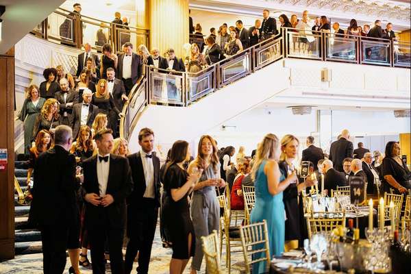 Many well-dressed people in formal attire gather at a grand event. Some walk on a large staircase, while others mingle near d