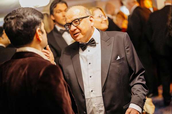 A distinguished man in a black tuxedo, bow tie, and glasses speaks intently to another man, whose back is to the viewer, at a