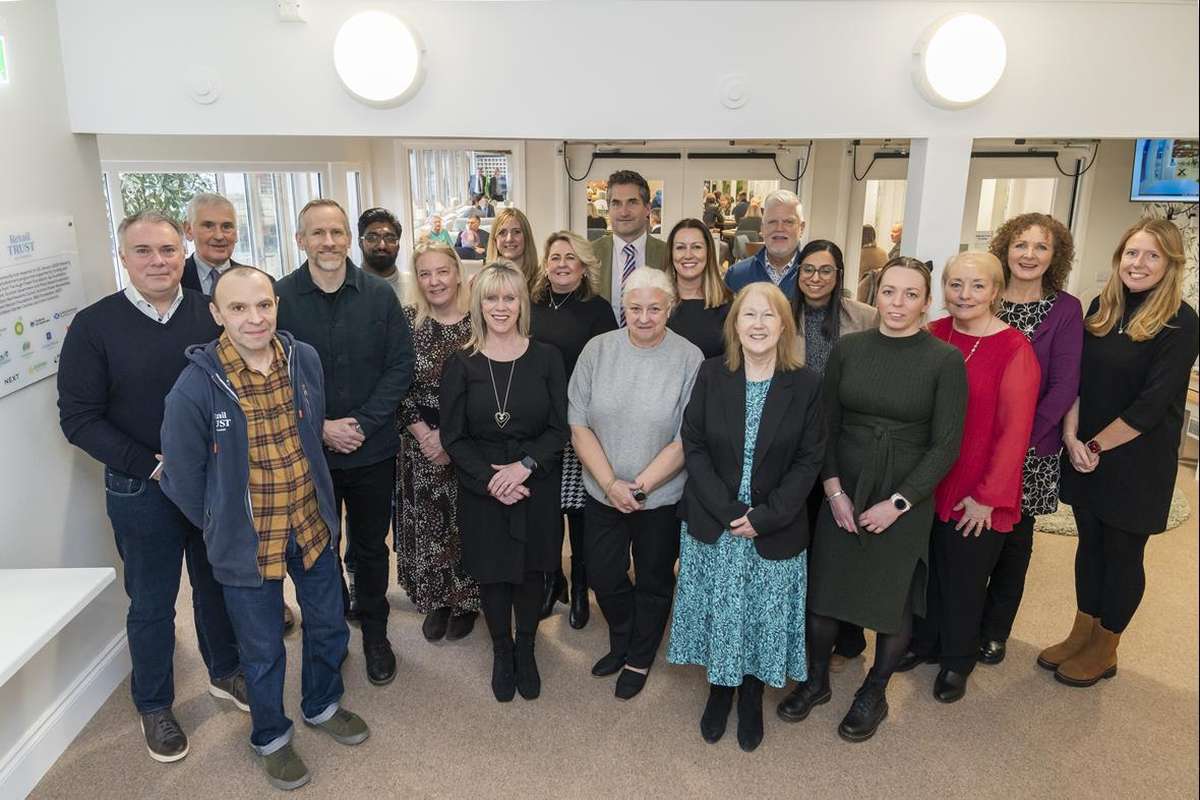 A diverse group of 20 smiling adults, men and women of various ages, pose for a group photo indoors in a professional setting.
