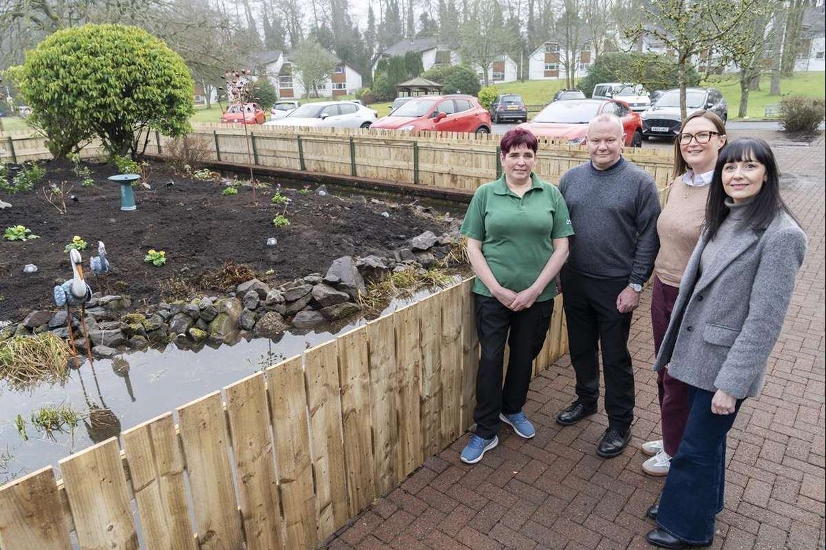Four people stand by a newly fenced garden area with a pond, small plants, and heron statues. Houses and cars are in the background.