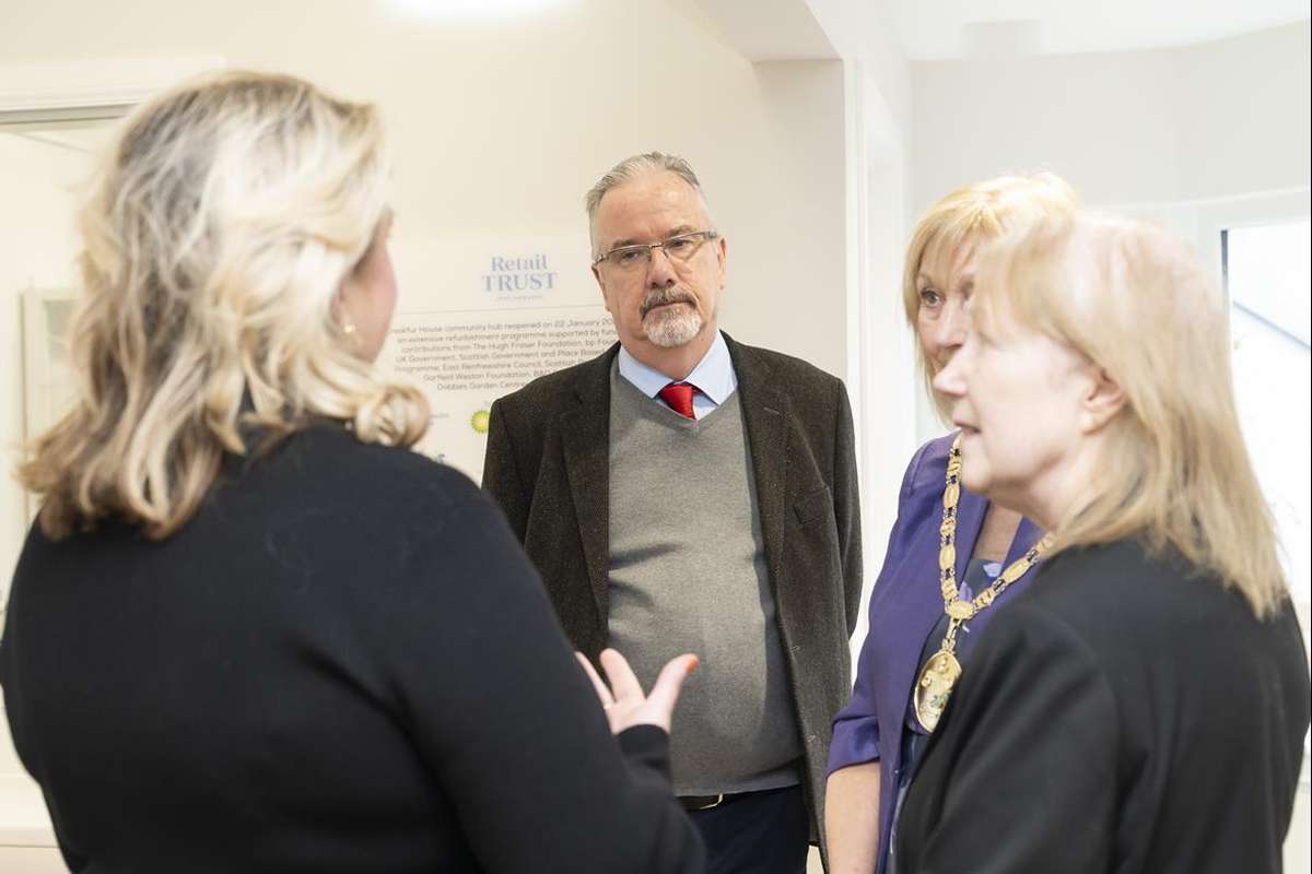 Four people in conversation indoors. A man in glasses and a red tie listens to a blonde woman gesturing, with two other women nearby.