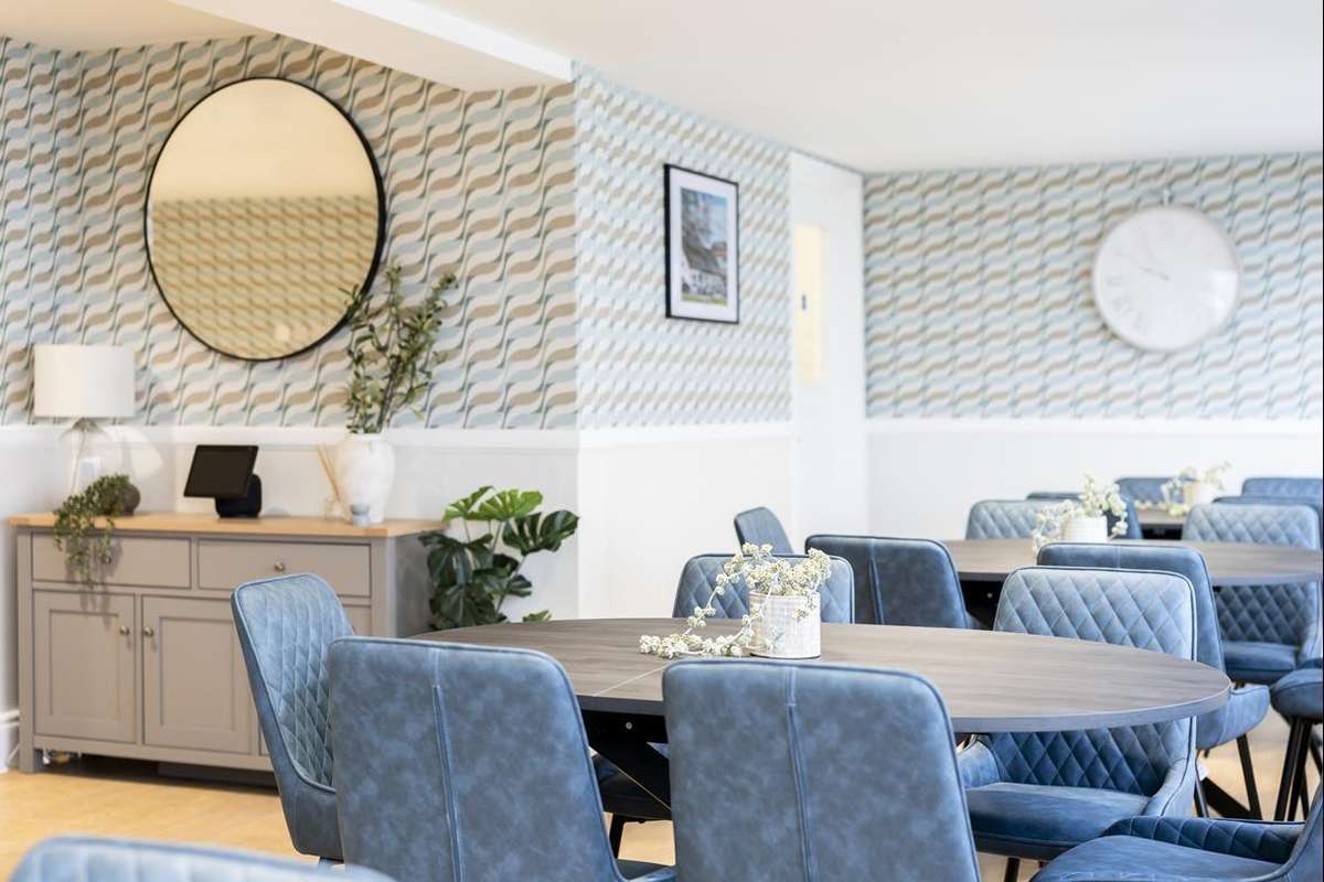 A modern common room with multiple round tables surrounded by blue upholstered chairs. Wavy blue and beige wallpaper covers the walls.