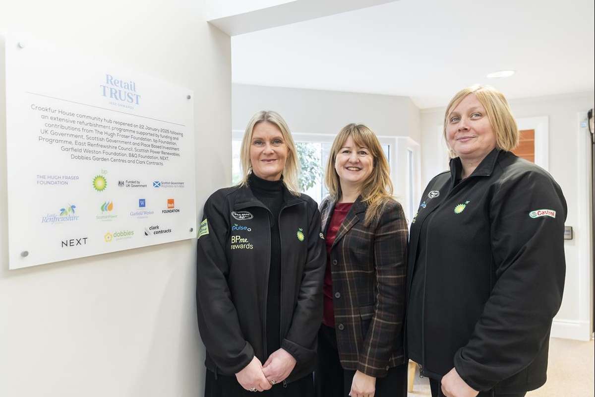 Three smiling women stand by a sign detailing the Retail Trust community hub's refurbishment. Two wear black logo jackets.