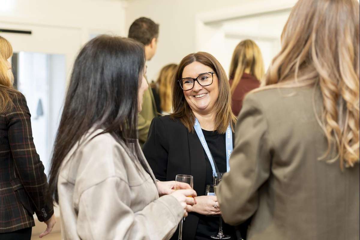 A cheerful woman in glasses and a black blazer with a lanyard talks to another woman, holding a drink at a busy indoor event.