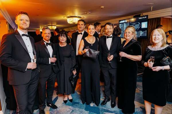 Eight smiling people in formal black attire hold champagne glasses at a glamorous indoor event with warm lighting and a table