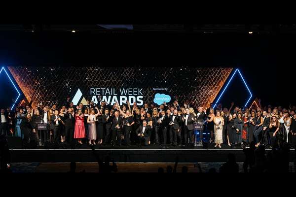 A large group of formally dressed people celebrate on stage at the Retail Week Awards, with a glowing screen displaying the e