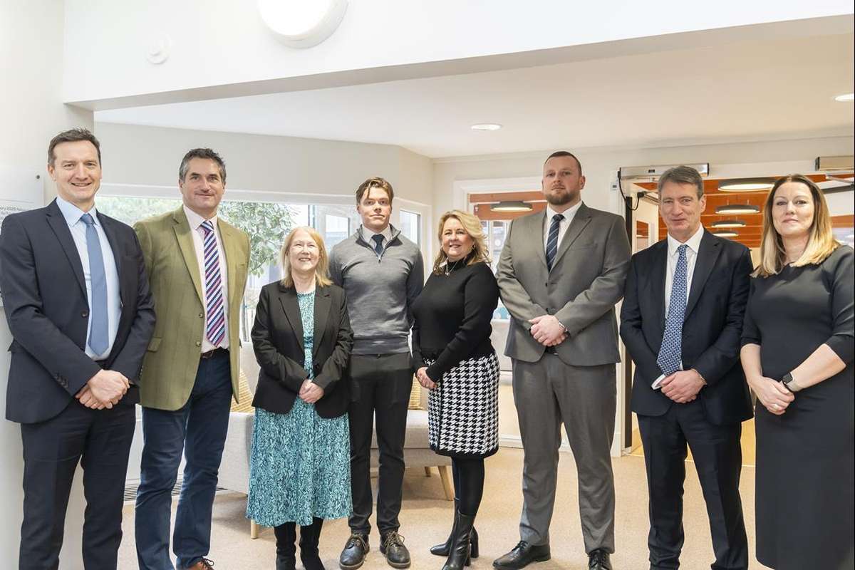 A group of eight professionals, five men and three women, dressed in business attire, stand smiling in a well-lit modern office or reception area.