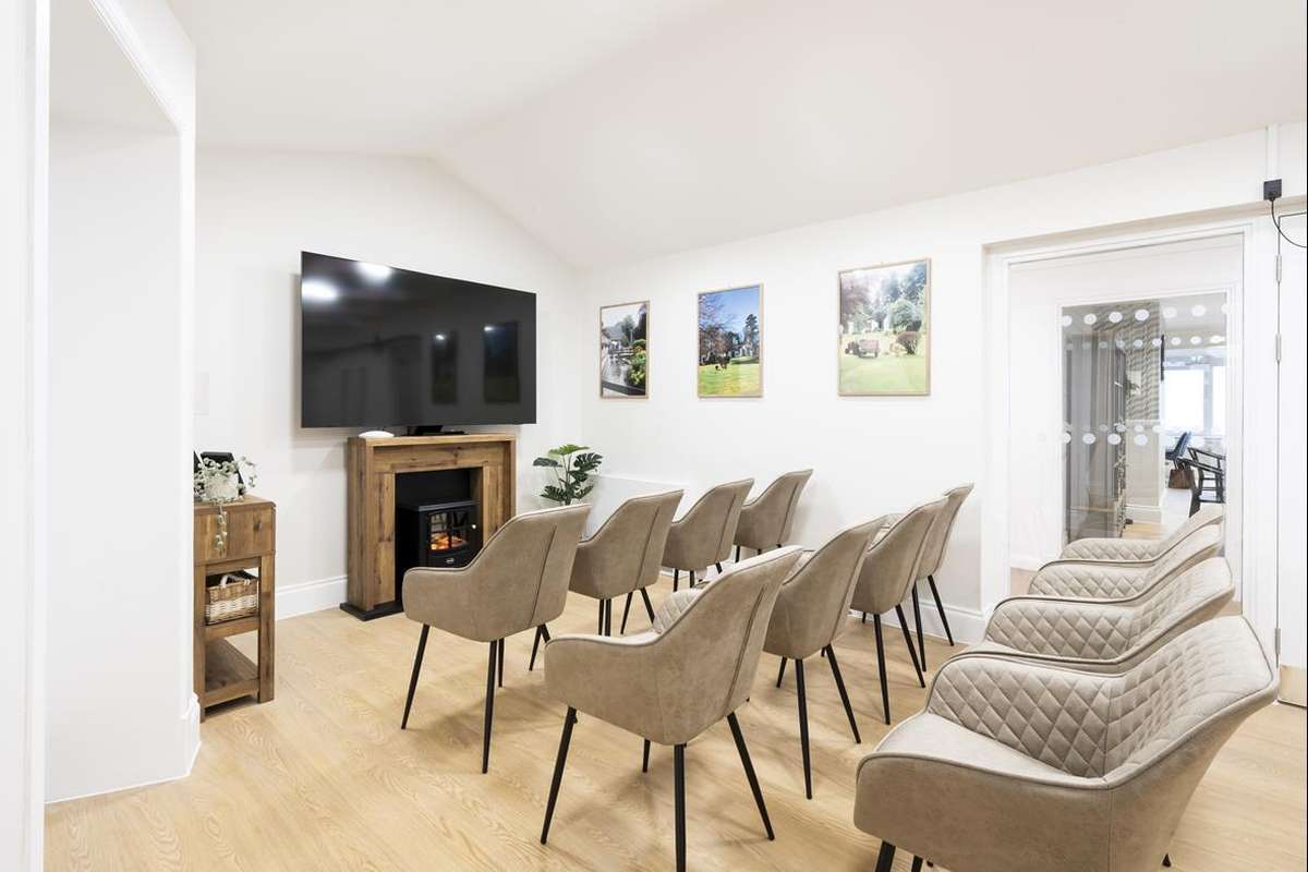 Modern room with rows of beige chairs facing a large TV above an electric fireplace. Three landscape pictures adorn the white wall.