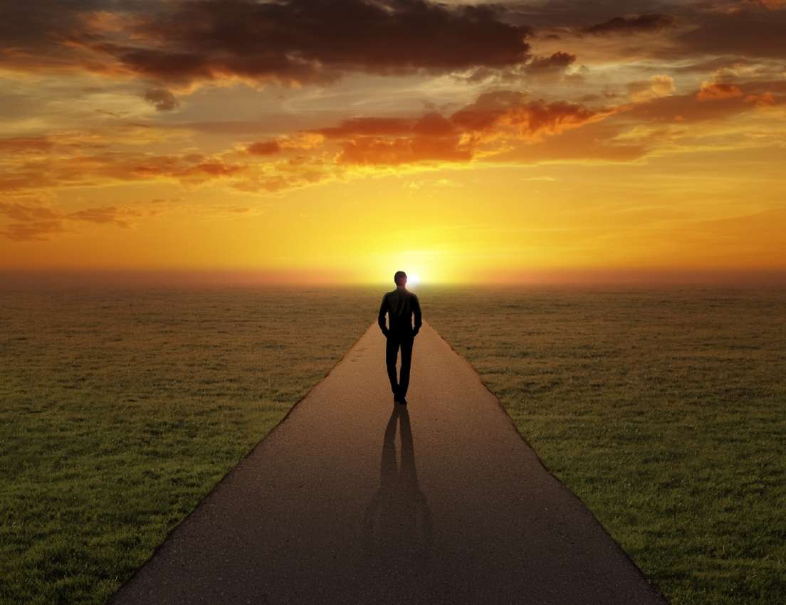 Silhouette of a person walking on a path towards a vibrant orange sunset over a flat grassy field.