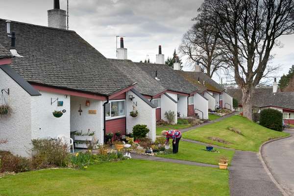 A woman tends a garden outside one of a row of white cottages with red trim, slate roofs, and green lawns under a cloudy sky.