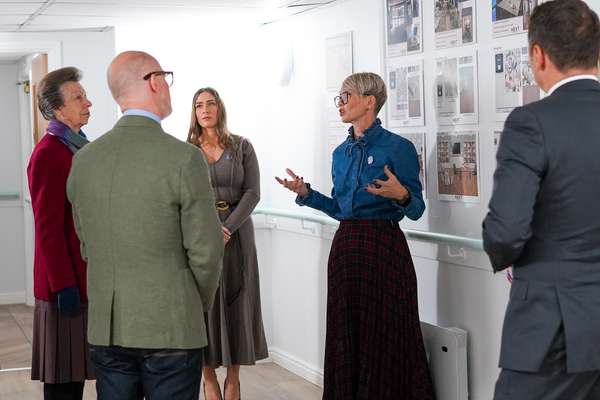 Princess Anne and a group listen to a woman presenting design boards on a white wall in a modern room.