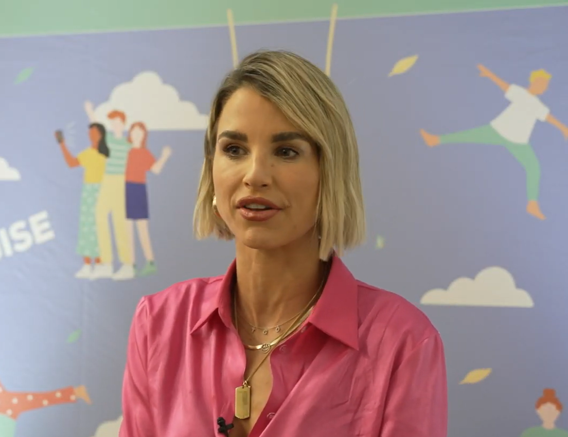 Blonde woman in pink shirt and gold necklaces speaking against a light blue wall with playful illustrations of people and clo