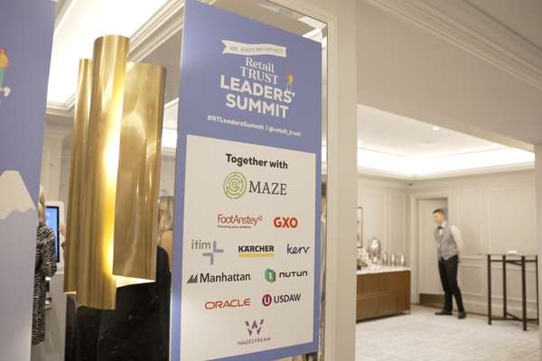 A blue and white sign for the "Retail Trust Leaders' Summit" with sponsor logos, in an elegant event venue with a server.