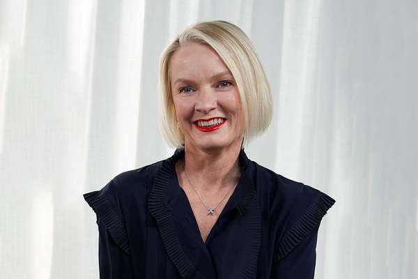A smiling woman with short blonde hair, blue eyes, and red lipstick, wearing a navy ruffled blouse and a silver necklace against a white background.