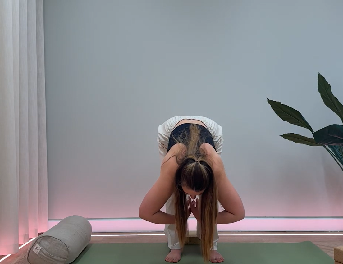 A woman in a black top and light pants performs a yoga forward fold on a green mat, hands clasped, in a modern studio.