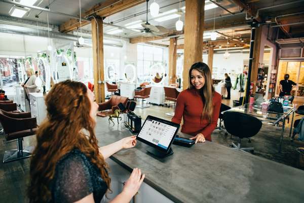 A woman in a red shirt helps a customer with long red hair at a tablet POS system in a modern salon.