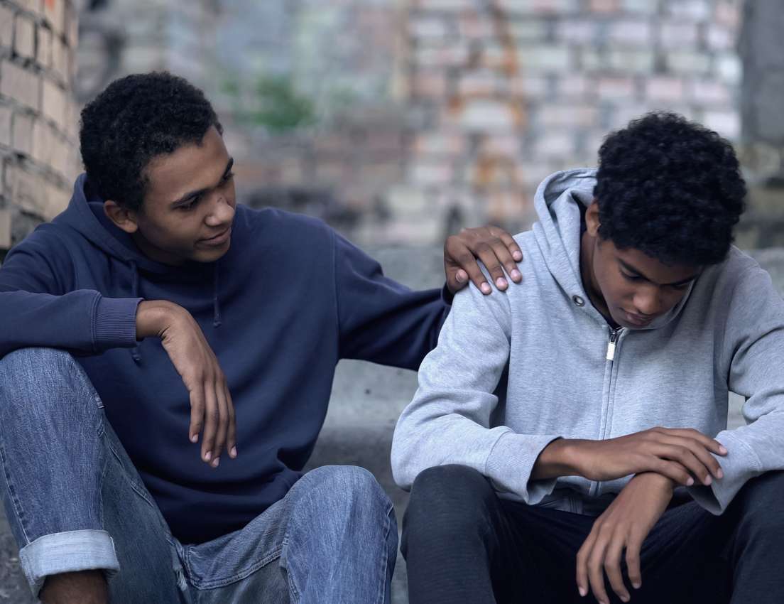 A young man in a blue hoodie comforts a sad, head-bowed young man in a grey hoodie while sitting on concrete steps.