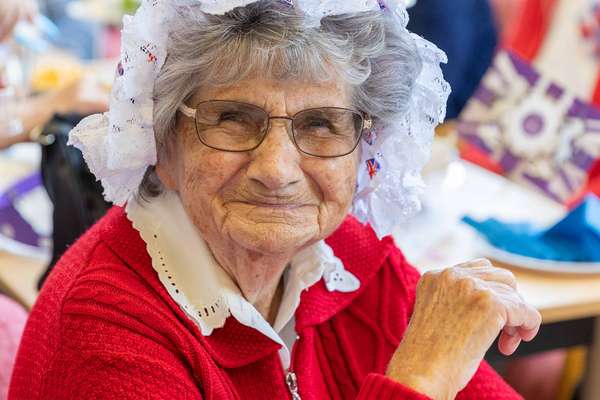 Smiling elderly woman with glasses, wearing a red cardigan and a white lace bonnet adorned with Union Jack flags.