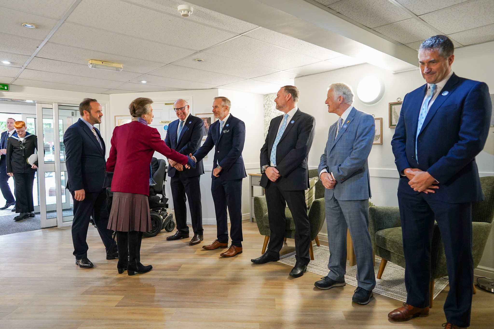 Princess Anne in a maroon jacket and brown skirt shakes hands with a man in a suit, surrounded by other men in suits in a room.