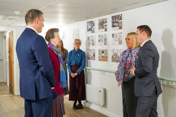 Princess Anne, in a red jacket and patterned scarf, speaks with a man in a suit and others in a hallway lined with interior d